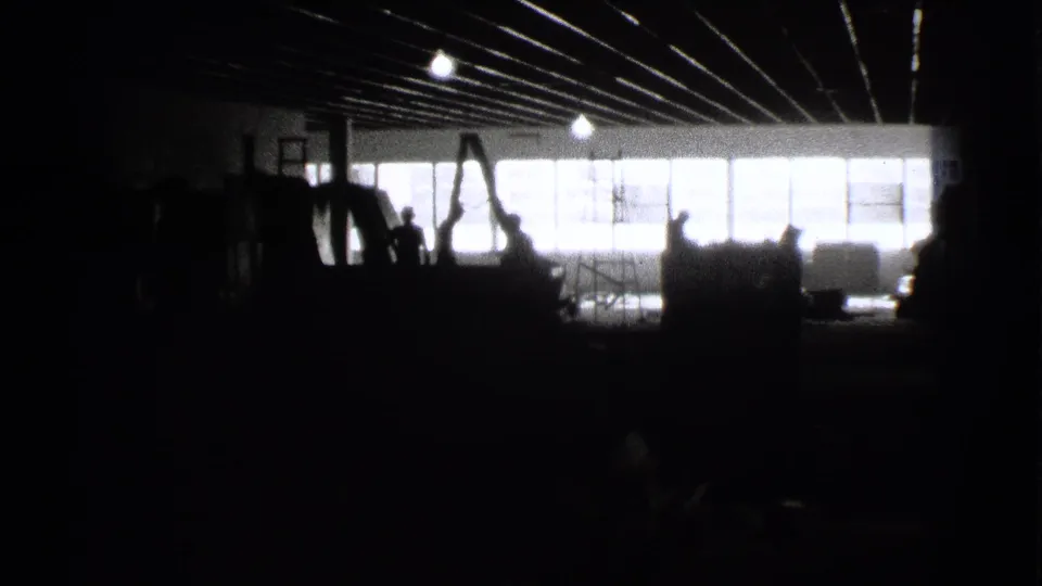 Black-and-white 8mm frame from 1939 Cleveland showing a dark industrial interior with backlit windows and people reduced to silhouettes.