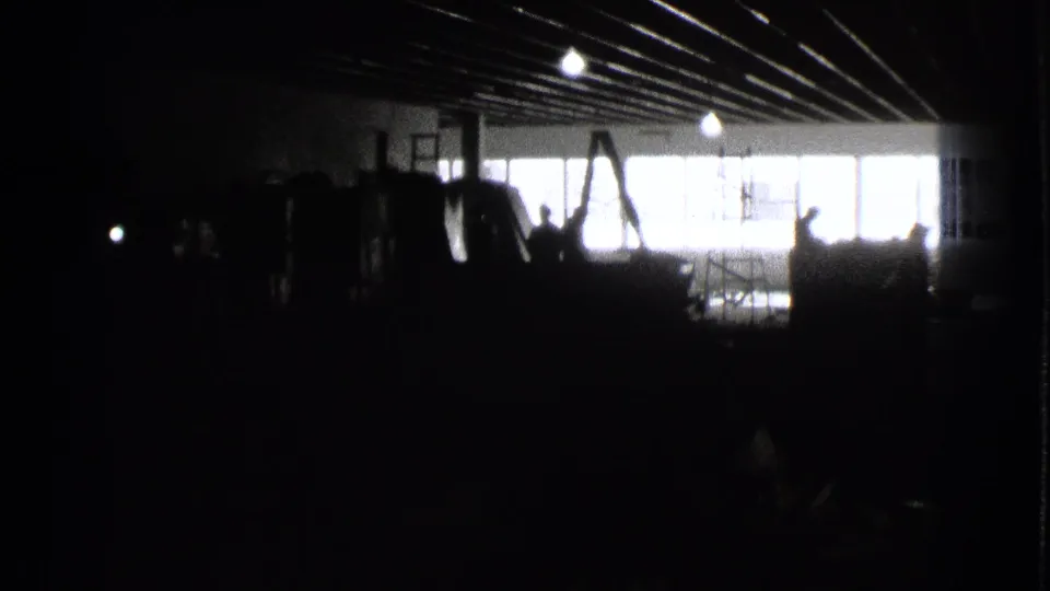 1939 Cleveland 8mm frame with a dark industrial-looking interior, bright horizontal window band, ceiling beams, and indistinct people.