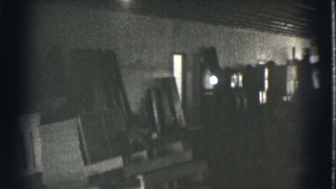 Dark interior with stacked objects, posts, ceiling beams, and a bright narrow opening in the background.