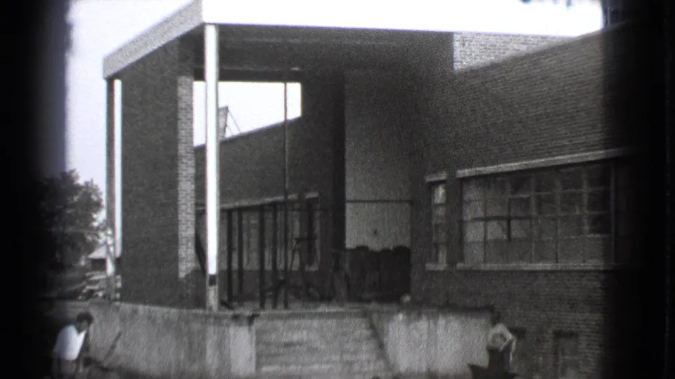 Final 1939 Cleveland frame with the brick building, covered entry, broad windows, exterior steps, and two people partly visible near the lower edge.