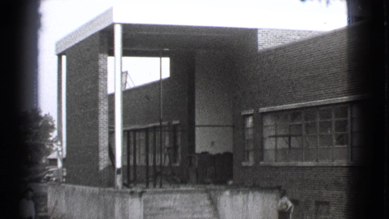 Brick building with covered entrance, columns, large windows, and small figures near the steps.