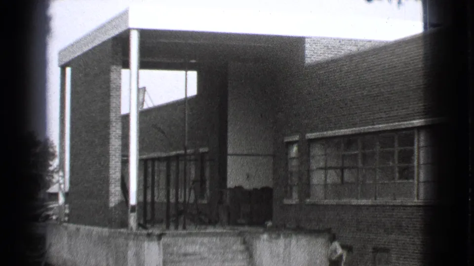 1939 Cleveland frame of a brick building exterior with a covered entry, slim columns, multi-paned windows, and a person partly visible near the lower left.