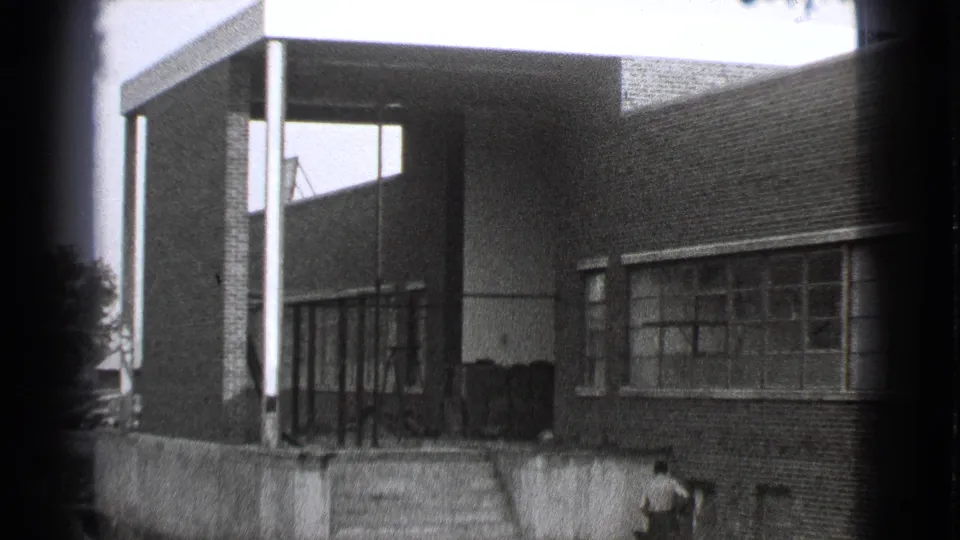 1939 Cleveland home-movie frame showing the building entrance, brick facade, window bands, steps or wall, and a person partly visible near the lower right.