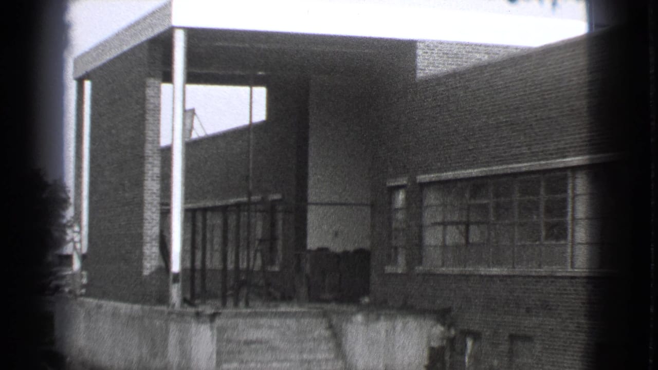 Brick factory entrance with covered steps and large windows in black-and-white footage.