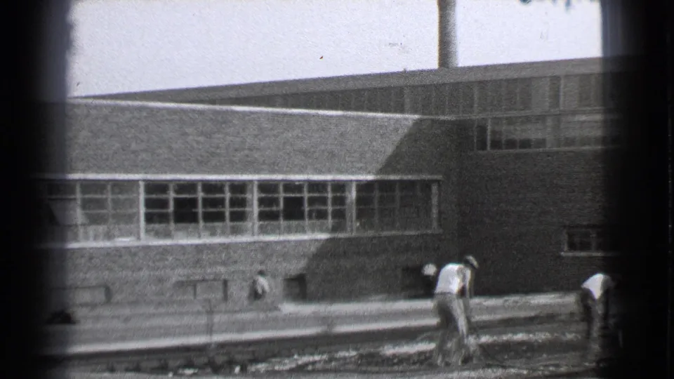 Black-and-white 8mm view of workers along railway tracks outside a Cleveland industrial building.