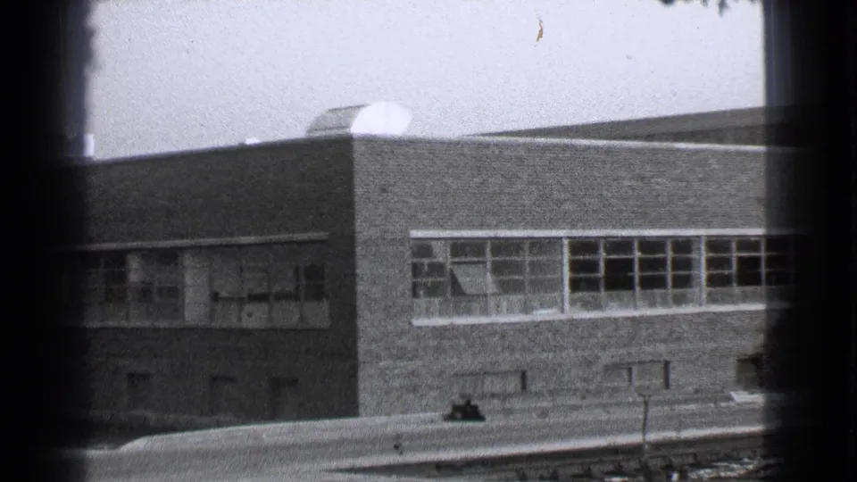1939 Cleveland frame of a low brick building with small windows and a rooftop vent, bordered by 8mm gate shadows.