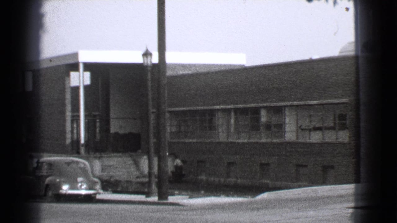Brick building beside a street with parked 1930s cars and a lamppost in front.
