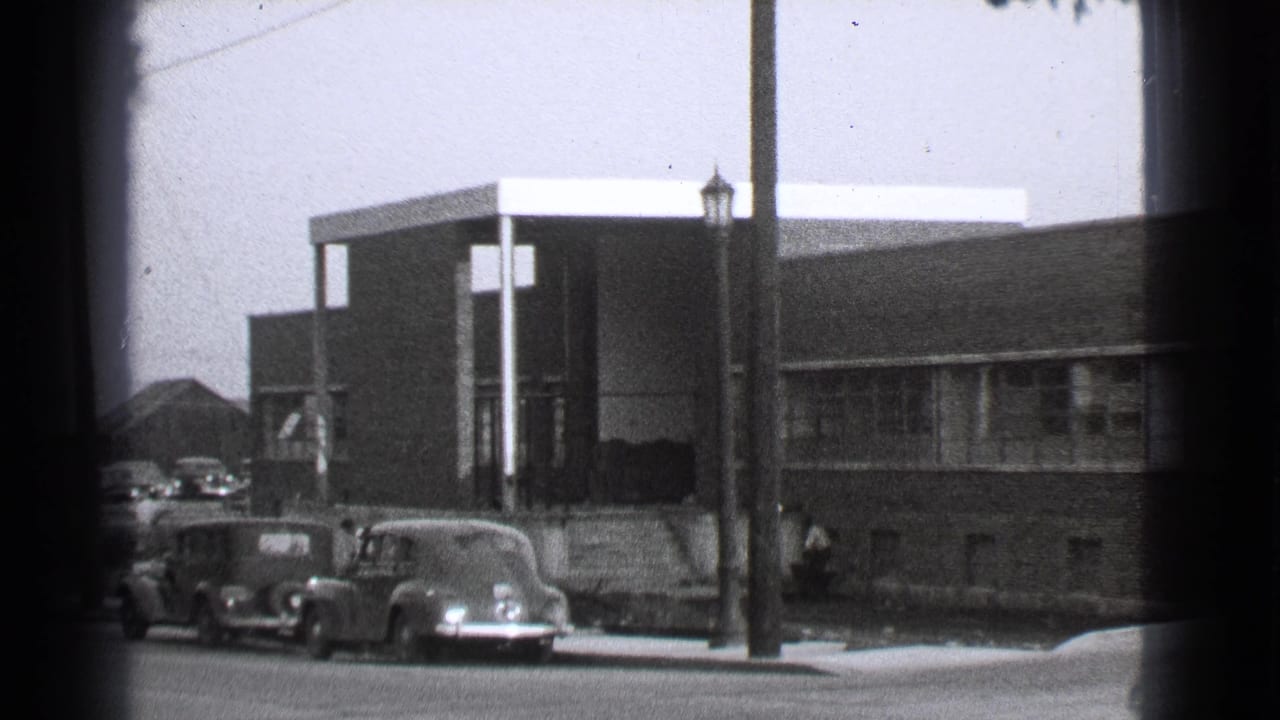 Parked classic cars line the curb in front of a brick building and covered entrance.