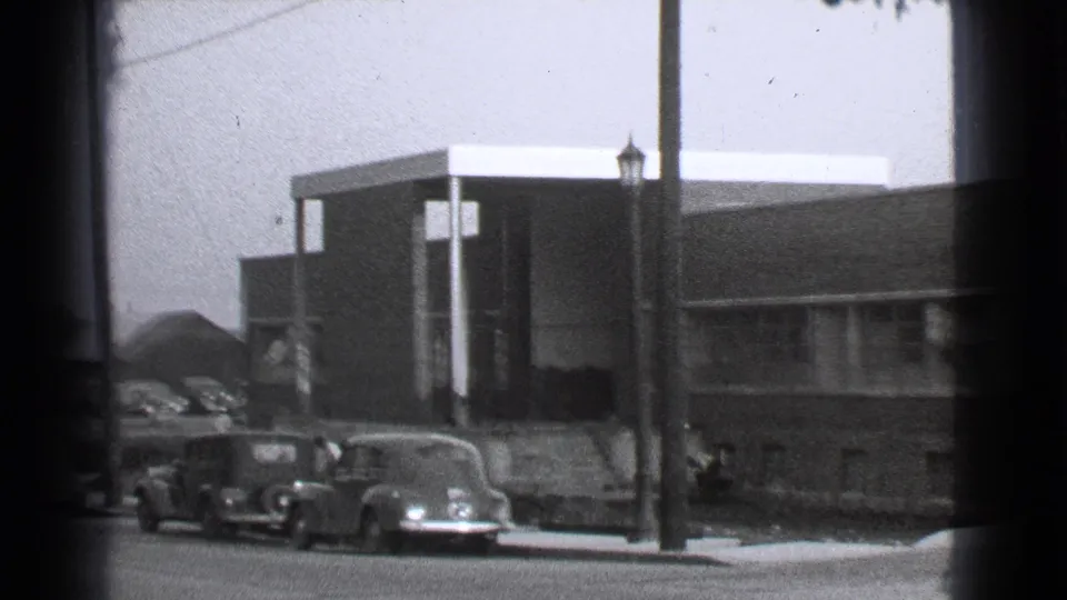 1939 Cleveland home-movie frame showing parked cars at a curb, a brick building with window bands, utility poles, and distant walking figures.