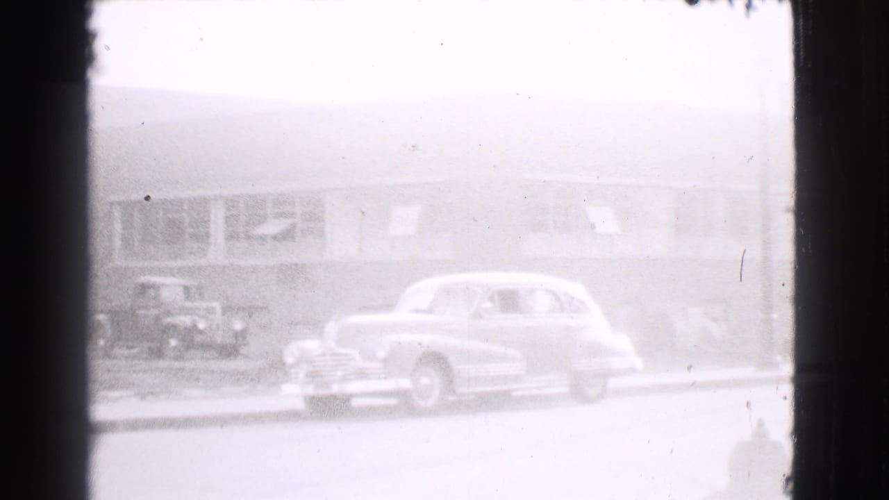 Curbside 1930s sedan in front of a low building with large windows.