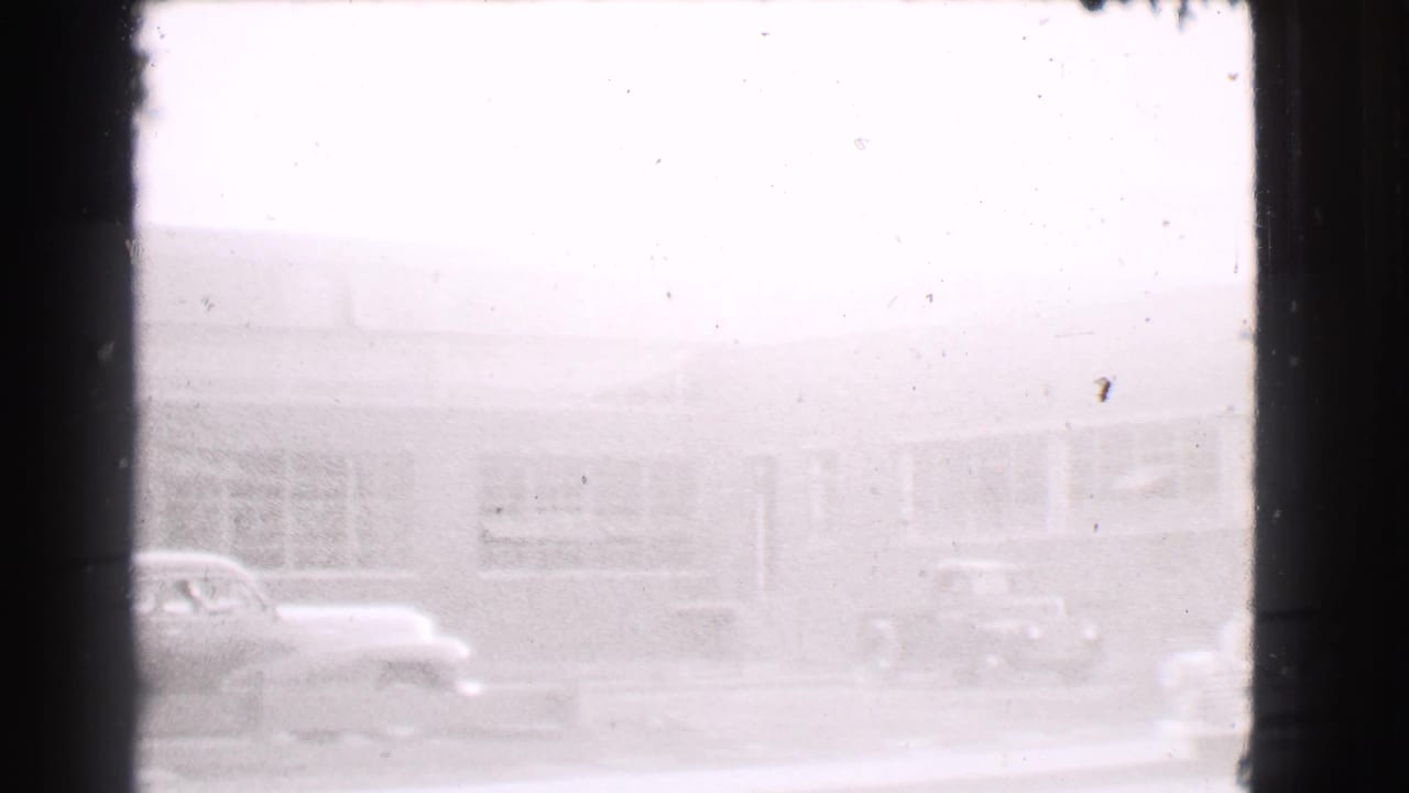 Pale street view through film frame edges with parked cars outside a low windowed building.