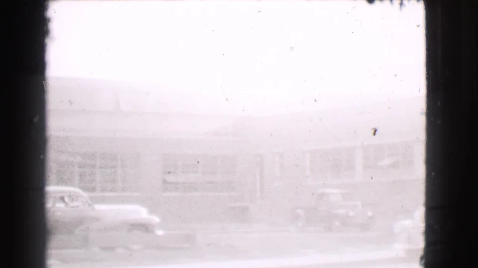 1939 Cleveland home-movie frame showing a bright street scene with cars parked along a low building with large windows.