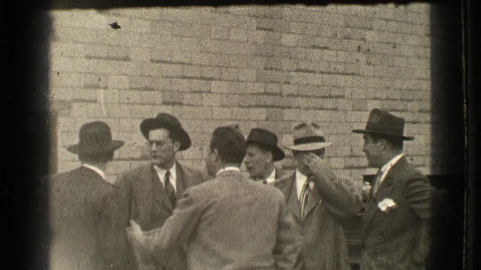 Group of men in suits and brimmed hats gather outdoors in 1939 Cleveland, talking near a brick wall.