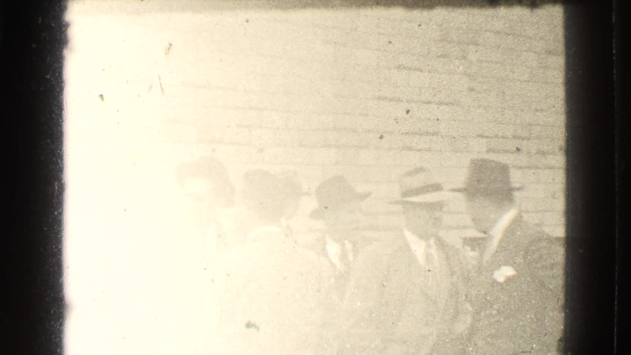 Overexposed frame of several men in suits and hats standing near a brick wall.