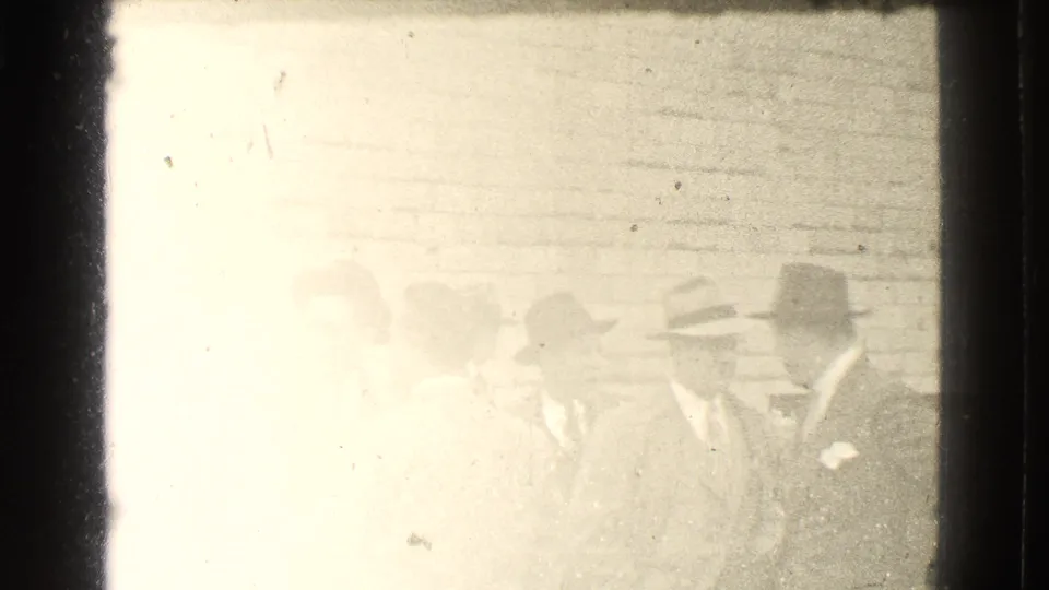1939 Cleveland home-movie frame showing adult men in suits and brimmed hats gathered outside a brick building, partly washed by exposure.
