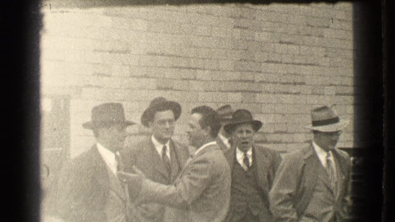 Men in suits and hats talk outside a brick building as one man gestures.