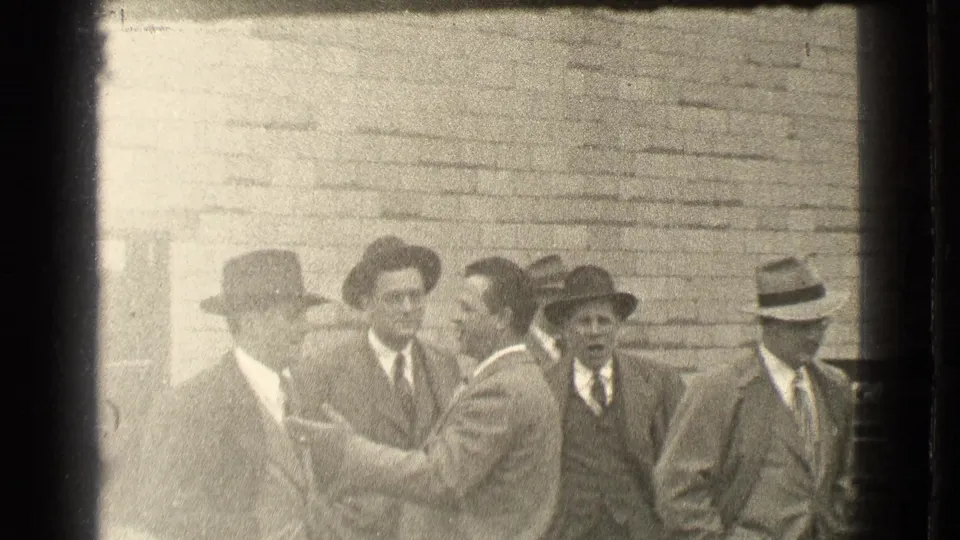 1939 Cleveland frame of suited adult men outside a brick wall, with one man turned sideways and gesturing during conversation.