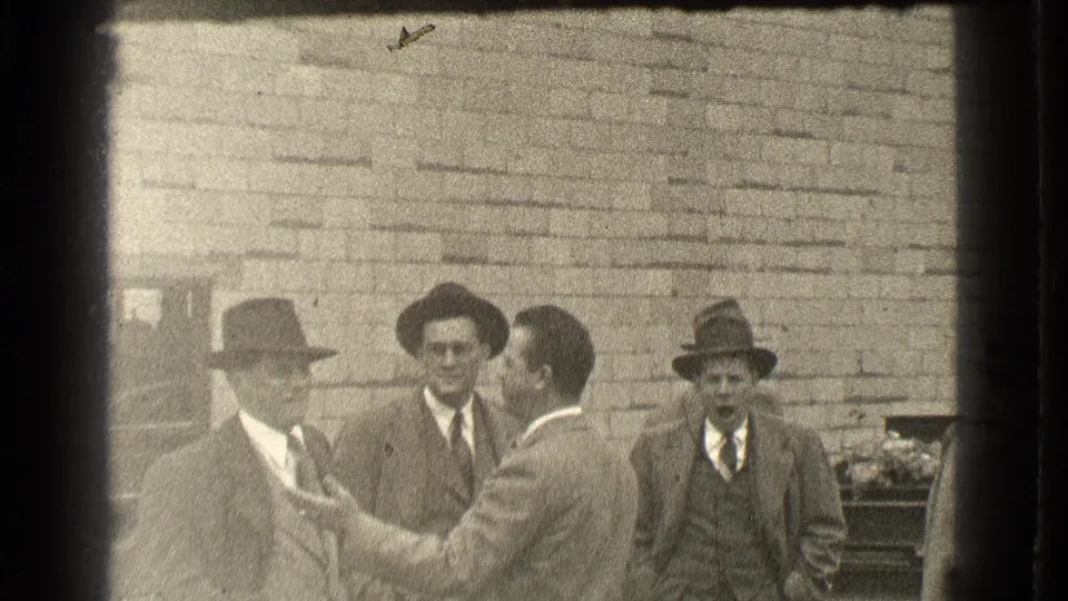 1939 Cleveland home-movie frame showing four suited men near a brick wall, including one gesturing and another facing the camera.