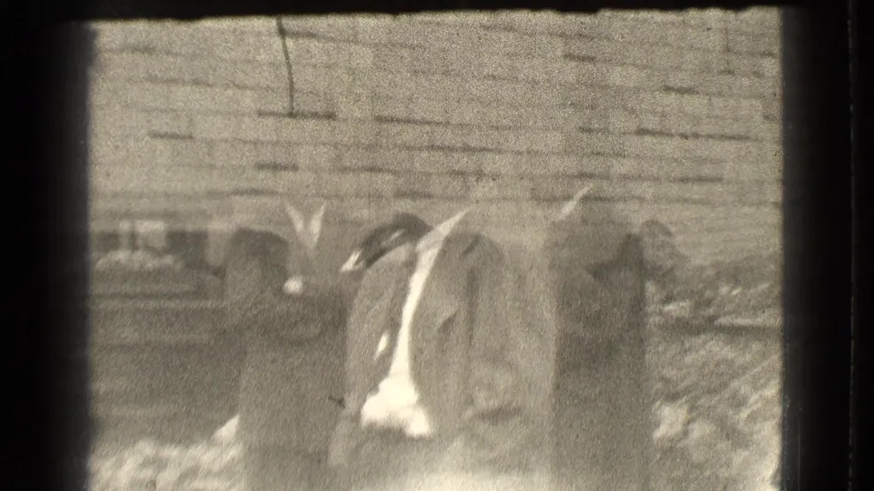 Blurred 1939 Cleveland frame with suited men partly obscured by motion or exposure near a brick wall and uneven outdoor ground.