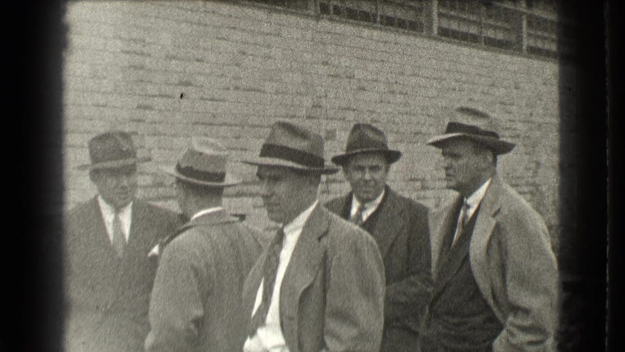Five men in suits and brimmed hats stand near a brick wall.