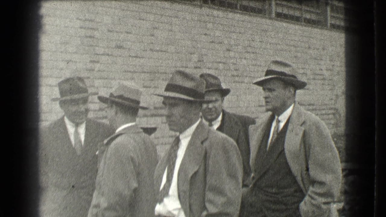 Men in suits and hats stand in a small group by a brick building.