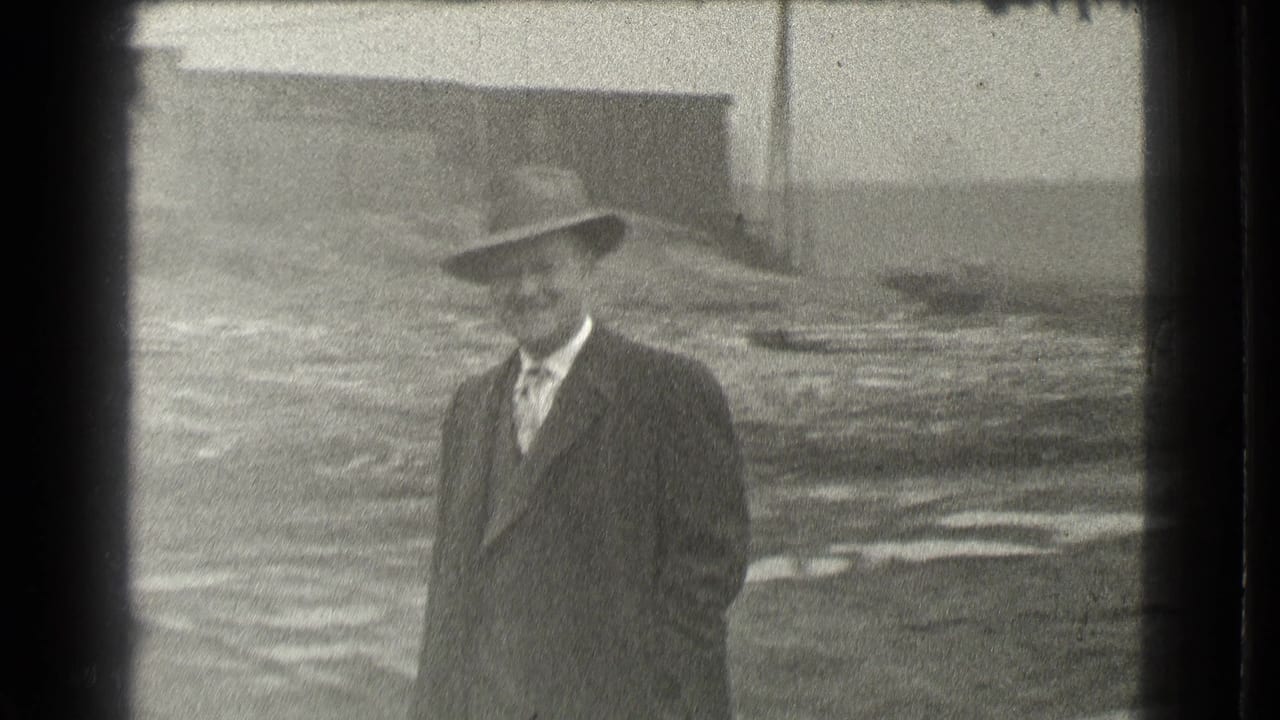 Man in a suit and fedora standing on unpaved ground near buildings.