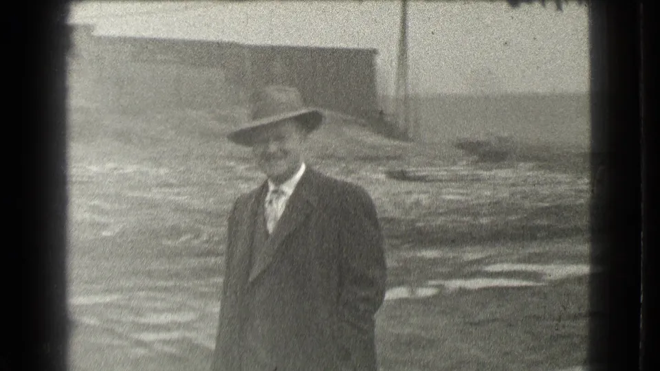 In 1939 Cleveland, a suited man in a fedora stands on rough ground near industrial-looking buildings and smiles toward the camera.
