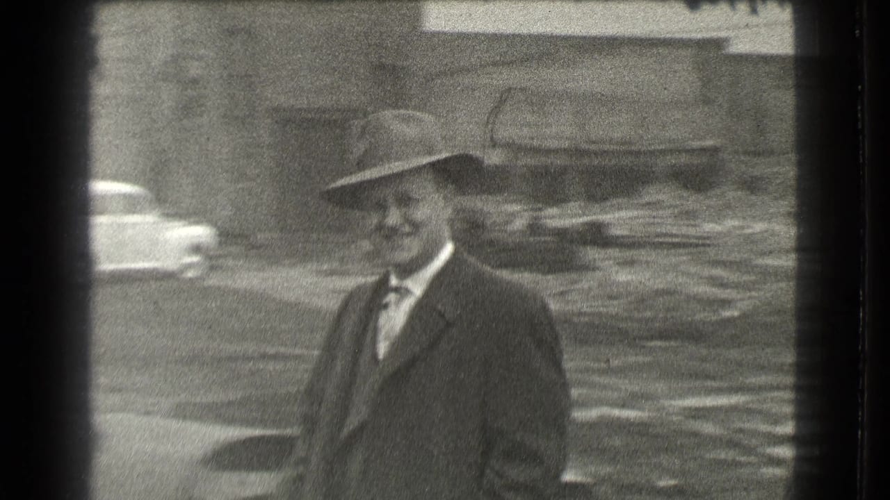 Man in suit and fedora standing near unpaved ground with a car behind him.