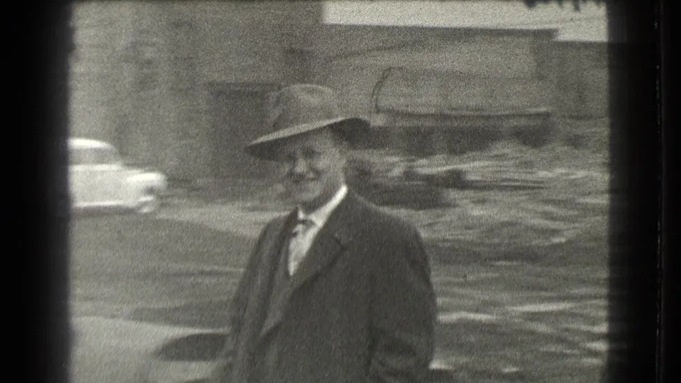 A suited man in a fedora stands in a Cleveland street-side industrial setting, with a rounded period car visible behind him.