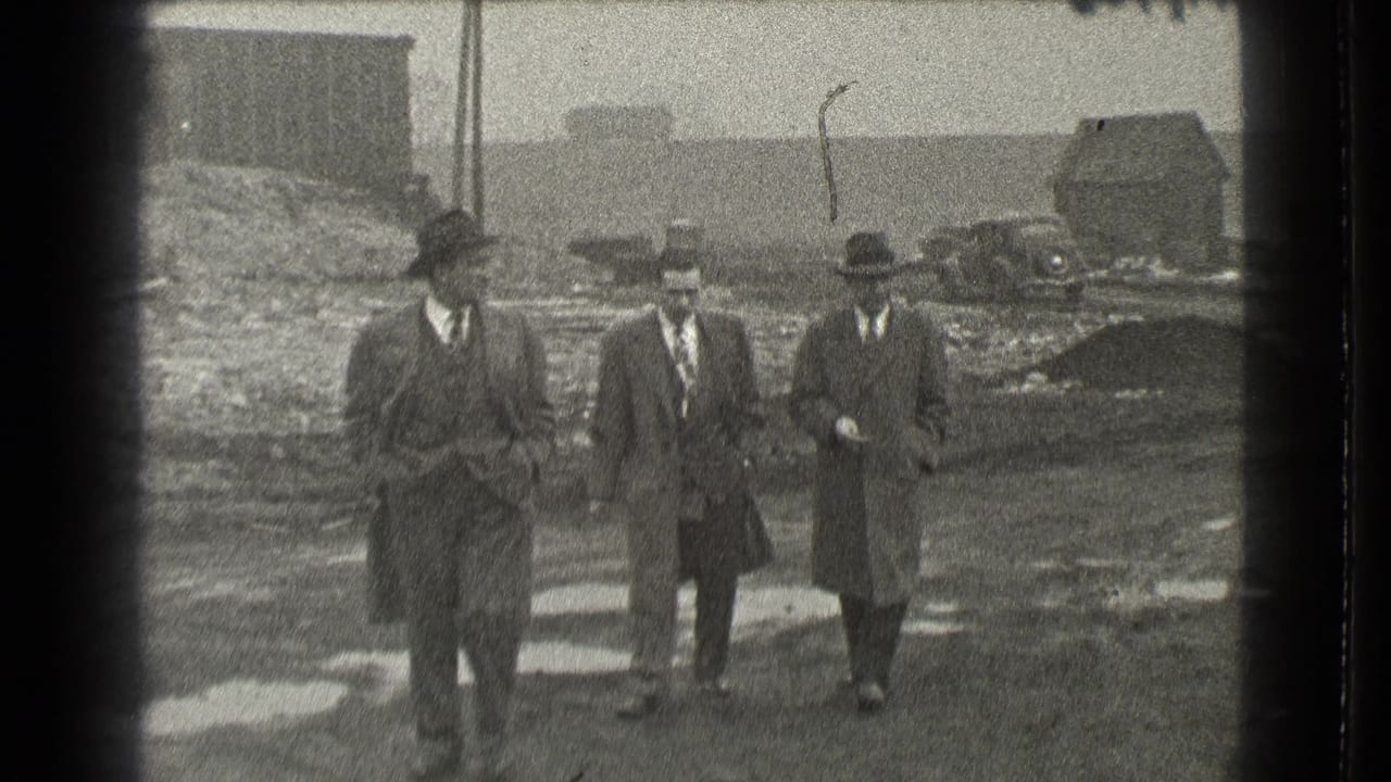 Three men in suits and hats walk across muddy ground toward the camera.