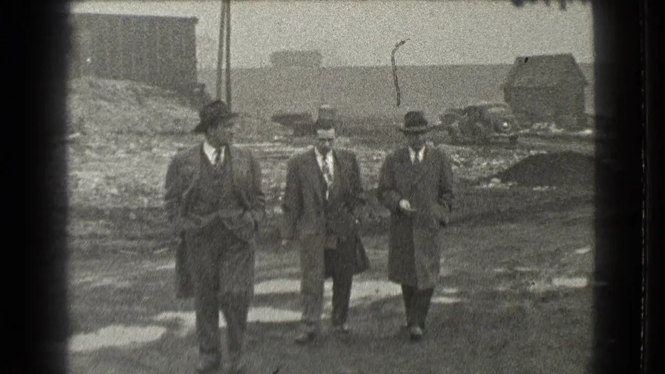 Three formally dressed men walk toward the camera at a rough outdoor site in 1939 Cleveland, with a small building and car behind them.