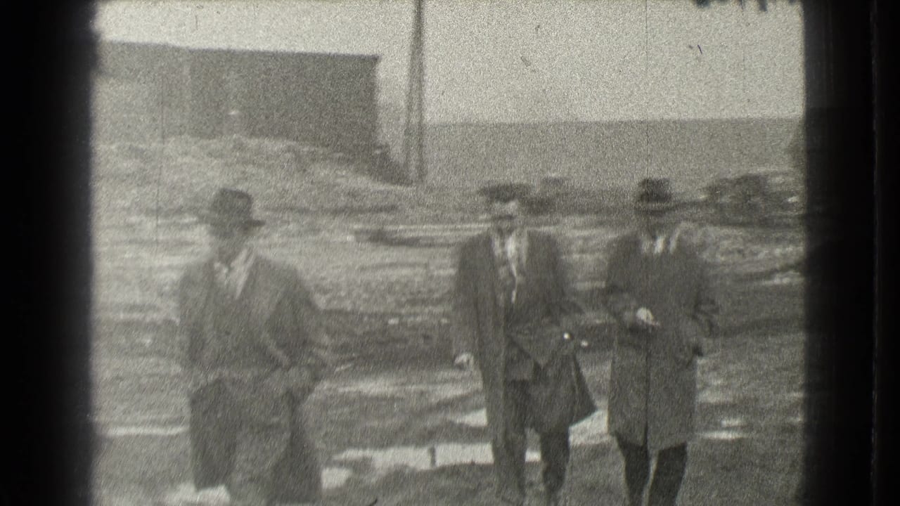 Three men continue walking toward the camera across an open muddy area.