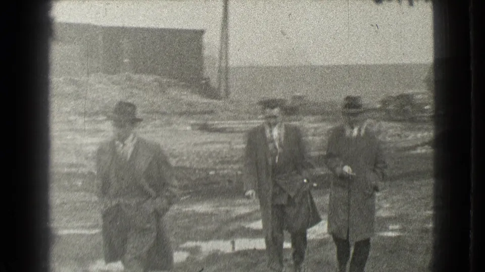 Three adult men in coats and hats move closer to the handheld camera in 1939 Cleveland, with rough ground and low structures behind them.