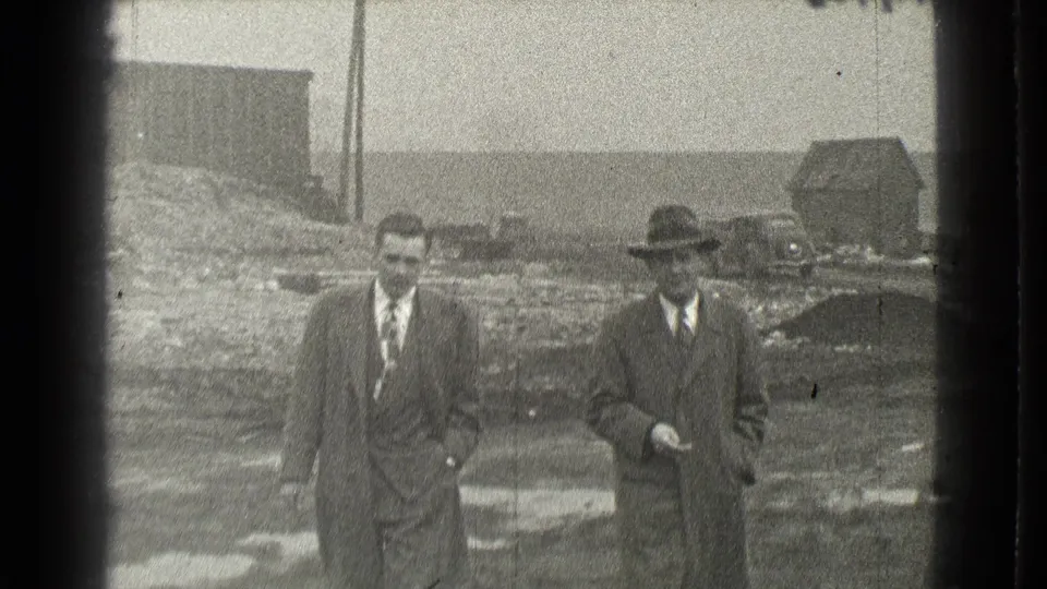 Two men in formal clothes pass close to the camera in 1939 Cleveland, with muddy ground, a utility pole, and a small building in the background.