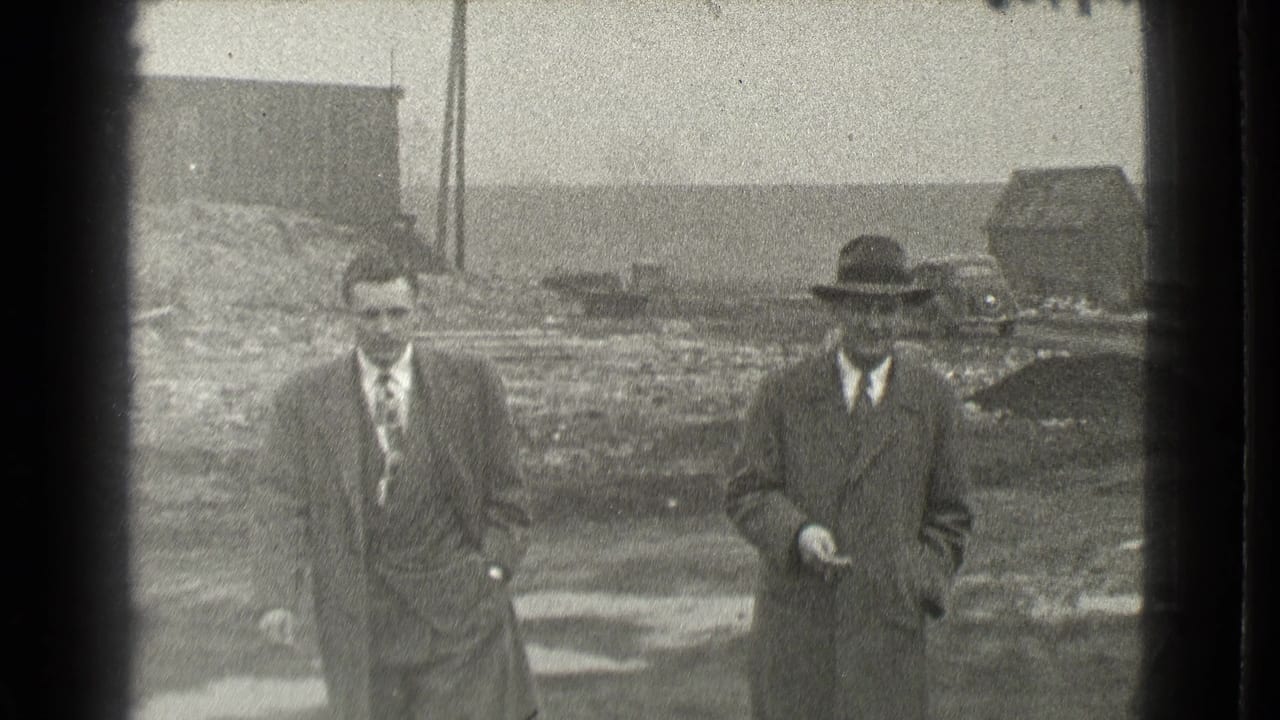 Two men in suits and hats walk across a muddy open lot.