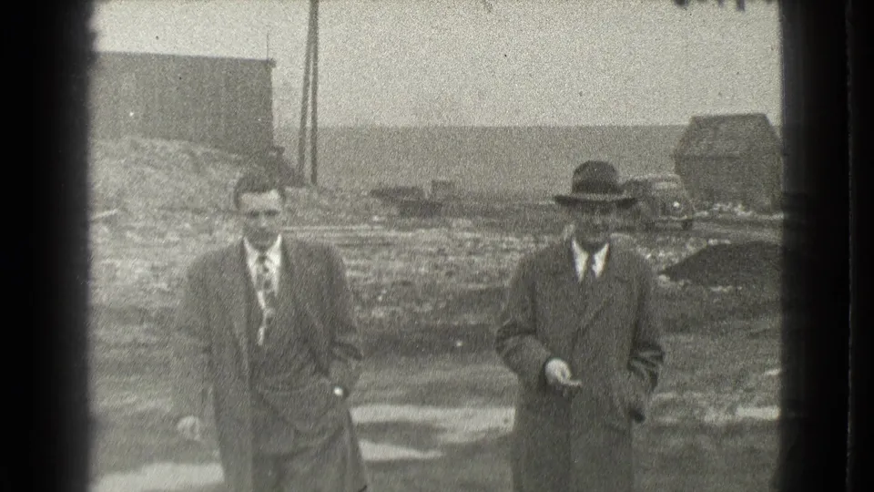 Two adult men in suits and brimmed hats walk together on a muddy lot in Cleveland, Ohio, in 1939 home-movie footage.