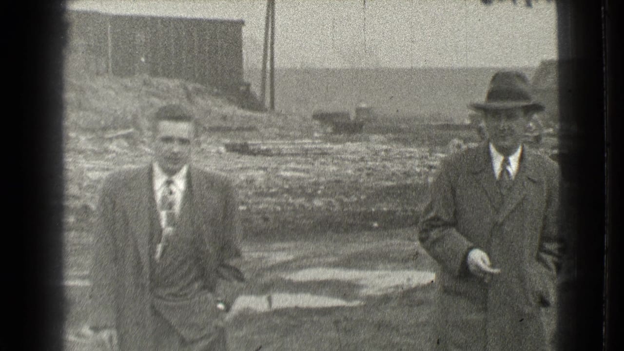 Two men in formal clothing appear on a muddy Cleveland lot, one closer to camera and another in a hat at right.