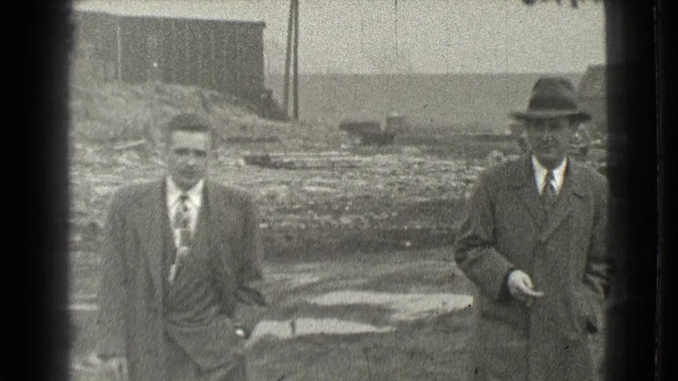 Two men in formal clothing appear on a muddy Cleveland lot, one closer to camera and another in a hat at right.