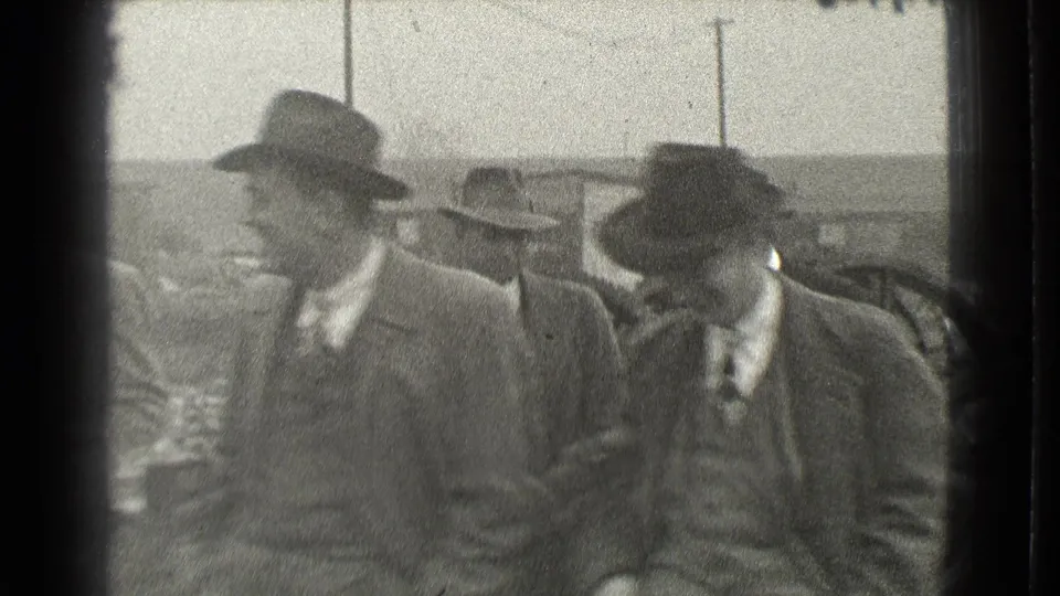 In 1939 Cleveland, several suited men in fedoras and overcoats walk together outdoors near industrial machinery and utility poles.