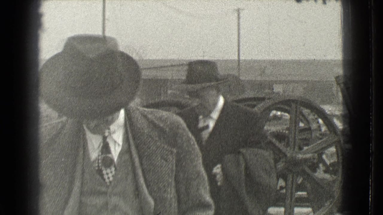 Man in a fedora looks down as other men and large wheels appear behind him.