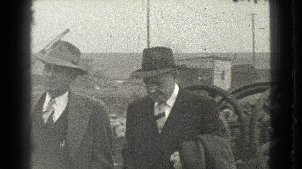 Two adult men in hats, suits and coats appear in the Cleveland outdoor industrial setting, with a large wheel behind them.