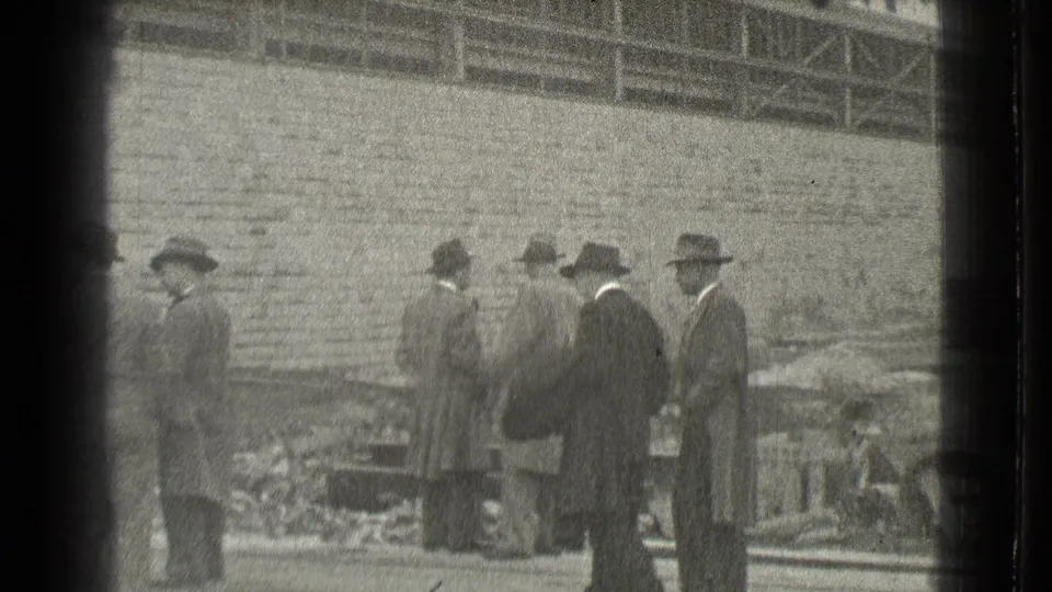 In 1939 Cleveland footage, several men in suits and brimmed hats gather beside a brick wall and scattered worksite debris.