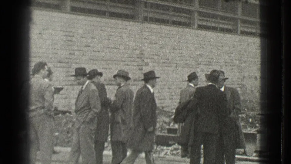 Suited men in hats congregate near construction debris in Cleveland, with a brick wall filling the background.