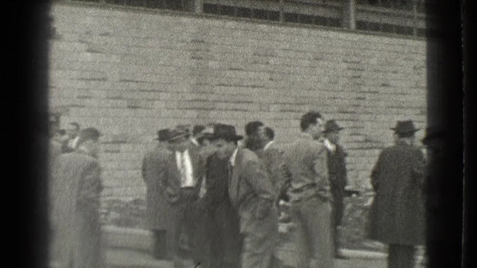 Men in period suits and hats remain gathered beside the brick wall and rubble in the 1939 Cleveland home-movie scene.