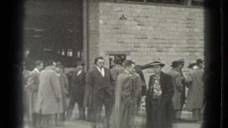 In Cleveland, Ohio in 1939, adult men in suits, overcoats, and hats appear to wait or talk outside a brick or stone building.