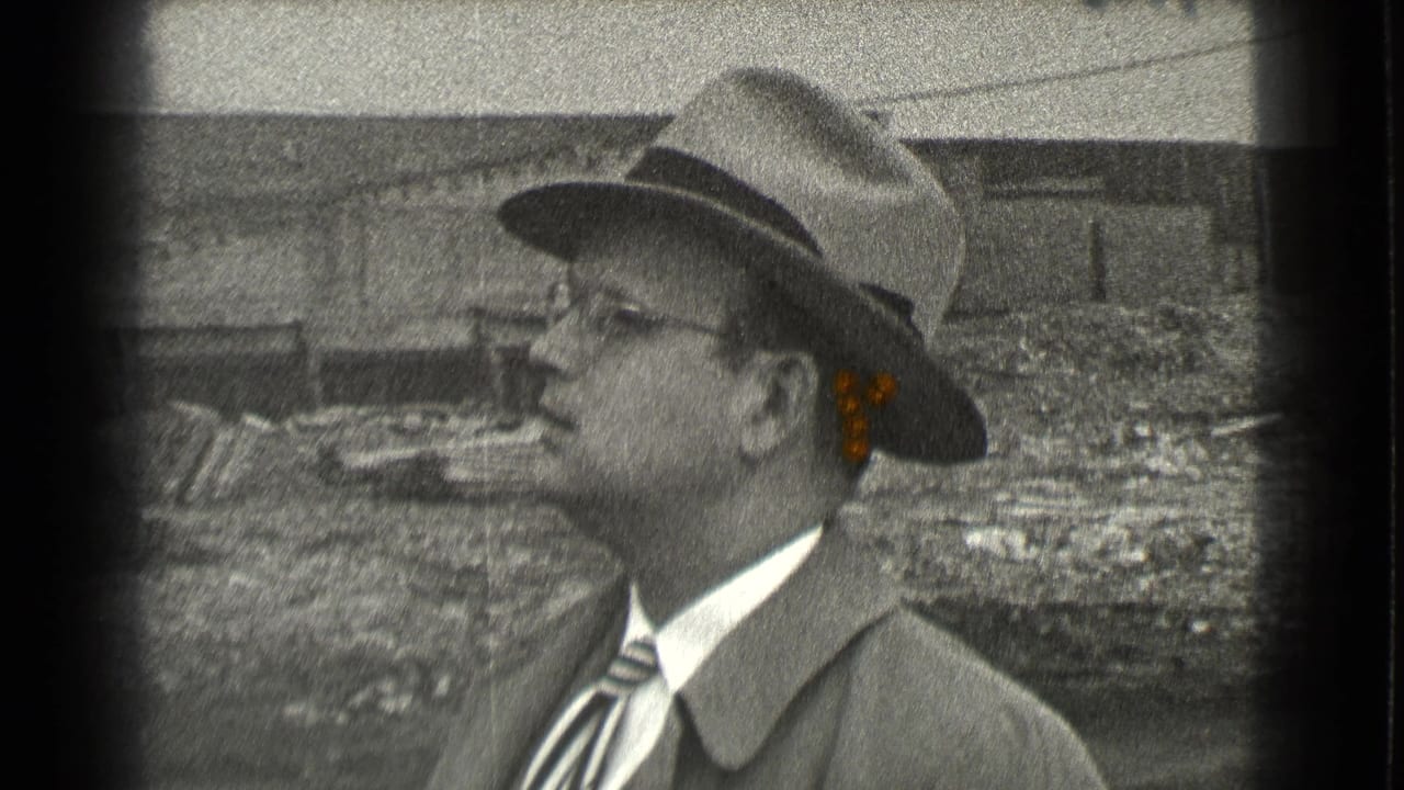 Man in a fedora and glasses smoking outdoors beside rough ground and a worn structure.