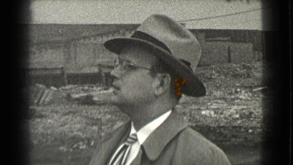 In 1939 Cleveland footage, a man in a fedora, glasses, suit jacket, and striped tie smokes outdoors near debris and a low worn building.