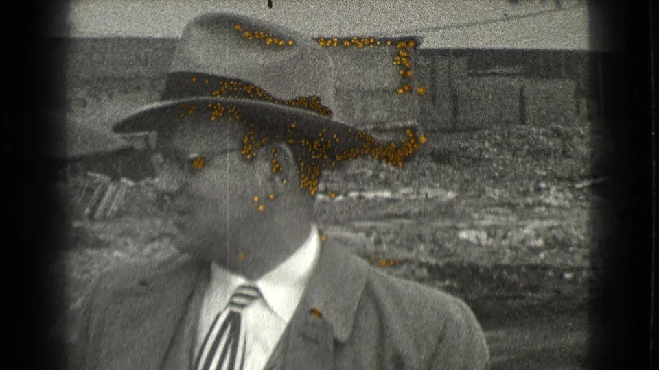 The 1939 Cleveland home-movie frame shows the man closer to camera, wearing glasses, a fedora, suit jacket, and striped tie.
