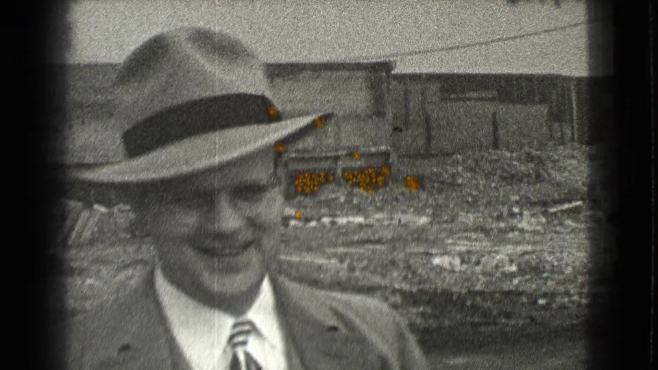 A suited man in a fedora appears to smile slightly while standing before rough ground and industrial buildings in Cleveland, Ohio in 1939.