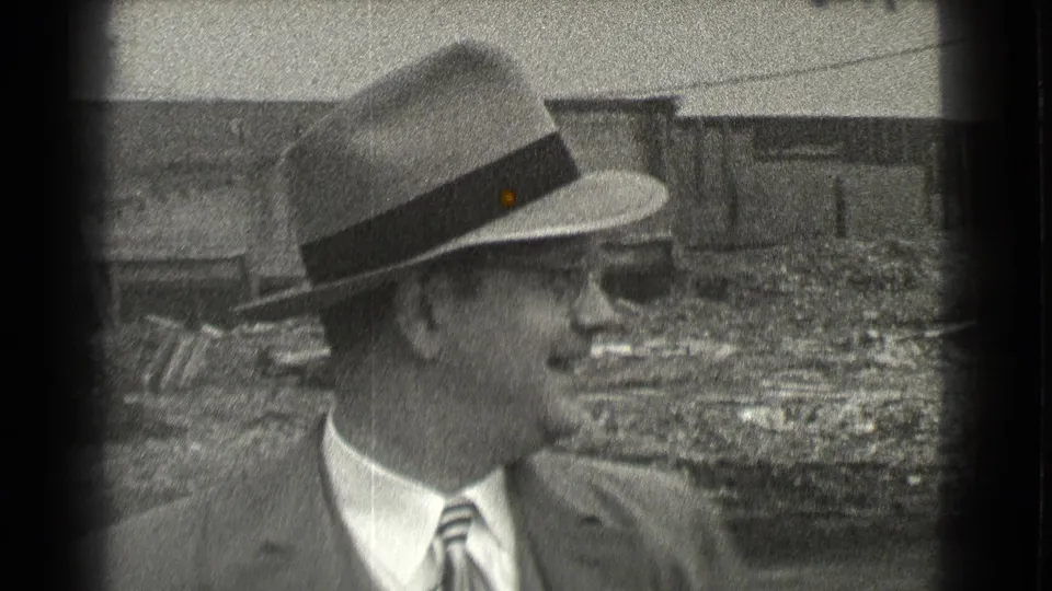 In Cleveland, Ohio in 1939, a suited man in a fedora and glasses faces sideways near debris and an unfinished building.